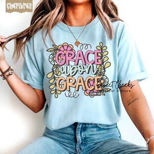 May include: Light blue t-shirt with the words "GRACE upon GRACE" in pink and orange, surrounded by floral and leaf designs. The text "JOHN 1:16" is below the main text. The shirt is a soft, casual style.