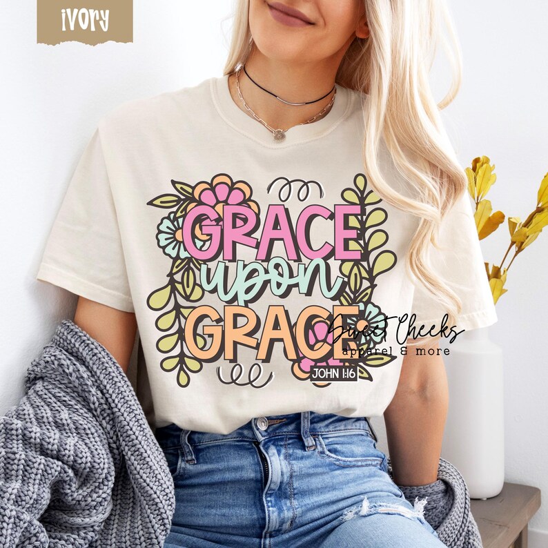 May include: Ivory-colored t-shirt with the words "GRACE UPON GRACE" in a colorful floral design. The text "JOHN 1:16" is below the main text. The shirt is a casual style.
