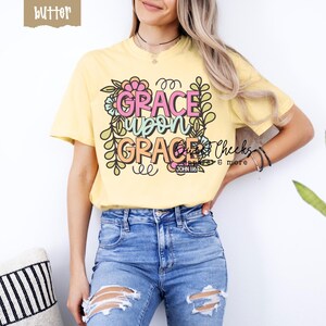 May include: Butter yellow t-shirt with the text "GRACE UPON GRACE" in pink and orange, surrounded by floral designs. The shirt also includes the text "JOHN 1:16". The model is wearing blue jeans.