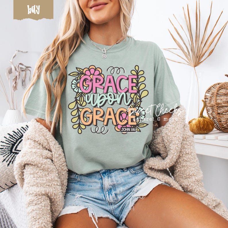 May include: Sage green t-shirt with the words "GRACE upon GRACE" in pink, orange, and yellow, surrounded by floral and leaf designs. The text "JOHN 1:16" is below the main text. The shirt is part of the apparel category.