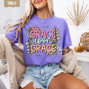 May include: A violet-colored t-shirt with the words "GRACE UPON GRACE" in a floral design. The text is in pink, orange, and white, with green and yellow floral accents. The shirt also has the text "JOHN 1:16".