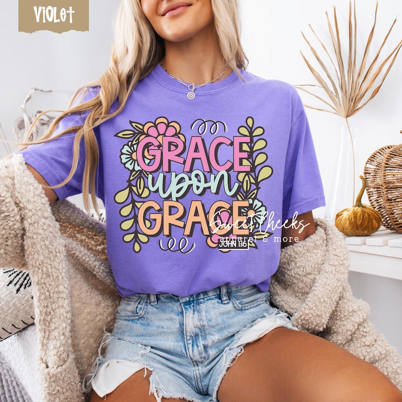 May include: A violet-colored t-shirt with the words "GRACE UPON GRACE" in a floral design. The text is in pink, orange, and white, with green and yellow floral accents. The shirt also has the text "JOHN 1:16".
