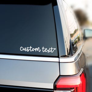 May include: A white vinyl decal applied to the rear window of a vehicle. The decal features the words custom text in a cursive script font, followed by a small heart icon. The sticker is positioned on the dark glass of the car window.