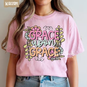 May include: A light pink t-shirt with the words "GRACE upon GRACE" in a floral design. The text is in pink, orange, and green. The shirt also has the text "JOHN 1:16".