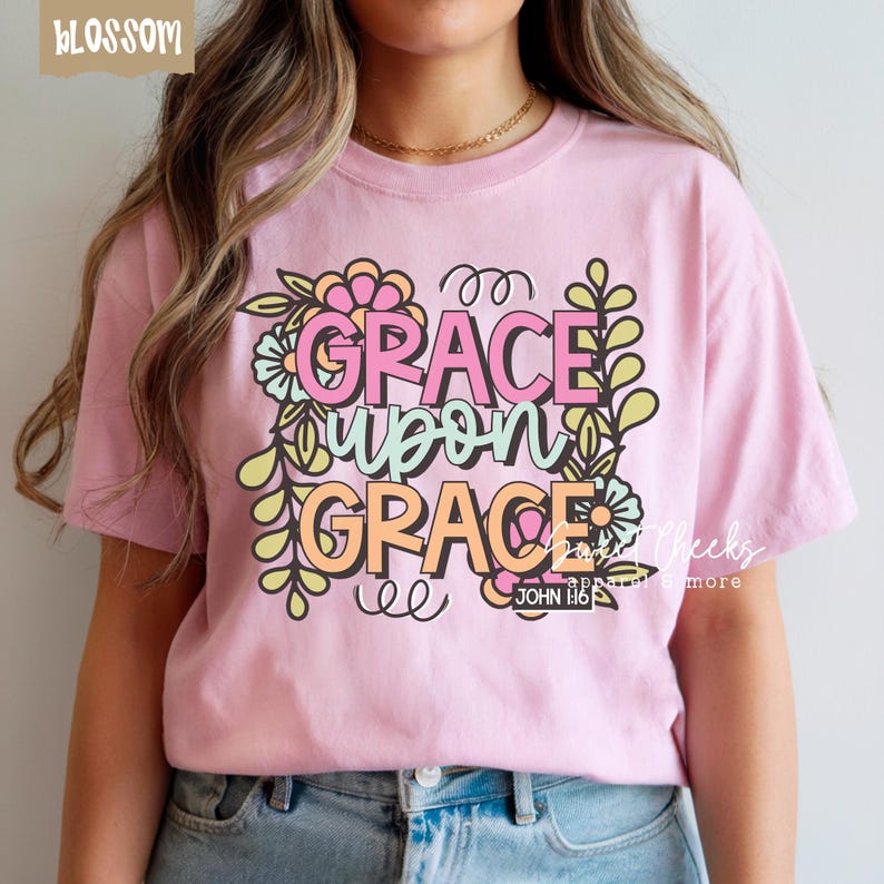May include: A light pink t-shirt with the words "GRACE upon GRACE" in a floral design. The text is in pink, orange, and green. The shirt also has the text "JOHN 1:16".