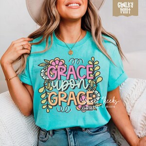 May include: Turquoise t-shirt with the words "GRACE upon GRACE" in pink, orange, and yellow, surrounded by floral and leaf designs. The text "JOHN 1:16" is below the words. The shirt has rolled-up sleeves.