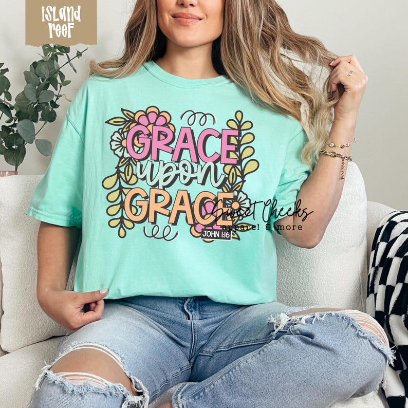 May include: A mint green t-shirt with the words "GRACE upon GRACE" in pink and orange, surrounded by floral and leaf designs. The text "JOHN 1:16" is at the bottom. The shirt is a comfortable, casual style.
