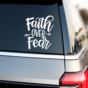 Może przedstawiać: Biała naklejka winylowa umieszczona na ciemnej szybie samochodu z napisem Faith OVER Fear w połączeniu czcionki pisanej i bezszeryfowej. Grafika strzałki z upierzonym ogonem przebiega poziomo przez środek napisu.