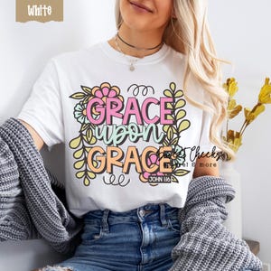May include: White t-shirt with the words "GRACE UPON GRACE" in pink and orange, surrounded by floral and leaf designs. The text "JOHN H:6" is below the main text. The shirt is part of the "Sweet Cheeks Apparel & more" collection.