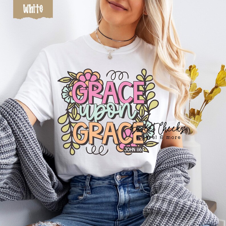 May include: White t-shirt with the words "GRACE UPON GRACE" in pink and orange, surrounded by floral and leaf designs. The text "JOHN H:6" is below the main text. The shirt is part of the "Sweet Cheeks Apparel & more" collection.