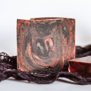 May include: Two bars of handmade soap with a black and red marbled design, sitting on a black lace fabric. A small orange pumpkin with a jack-o'-lantern face is in the background.