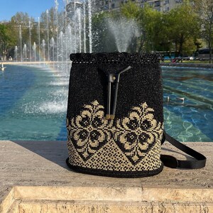 以下が含まれることがあります： 黒とベージュのクロシェの巾着バックパック。革のストラップと金のアクセントが付いています。バックパックは噴水の前、石の表面の上に置かれています。