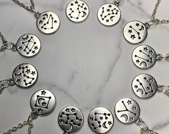 May include: A collection of silver-toned constellation necklaces. Each pendant features a different zodiac constellation design with stars and crescent moons. The necklaces are displayed on a light-colored surface.