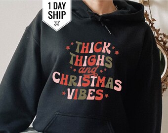 May include: Black hooded sweatshirt with the text "THICK THIGHS and CHRISTMAS VIBES" in a retro font. The sweatshirt has a drawstring hood and a front pocket. The text is in red, green, and pink colors. The model is wearing ripped blue jeans.