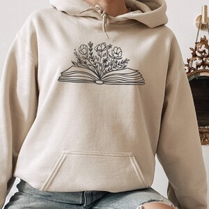 May include: A beige hoodie with a black outline of an open book with flowers growing out of it.