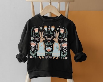 Könnte beinhalten: Schwarzer Pullover mit einem Blumendruck mit zwei Kaninchen in blauen Kleidern. Der Druck enthält Blumen, Blätter und geometrische Formen in den Farben Rosa, Blau, Grün und Braun.