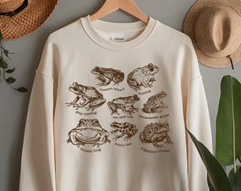 May include: Cream-colored sweatshirt featuring a brown illustration of various frog species, each labeled with its name. The design includes nine different frog types. Two straw hats hang on the wall behind the sweatshirt.