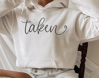 May include: A white hoodie with the word "taken" written in cursive with a heart symbol next to it. The model is wearing the hoodie with a pair of light blue denim shorts and white socks.