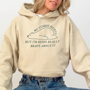 May include: A beige hoodie with a graphic of a bear lying down and the text "My tummy hurts but I'm being really brave about it".