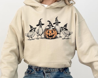 Könnte beinhalten: Ein beiger Hoodie mit einem Aufdruck von fünf Hühnern, die Hexenhüte tragen und eine ausgehöhlte Kürbislaterne halten. Die Hühner stehen vor einem Kürbis mit einem geschnitzten Gesicht.