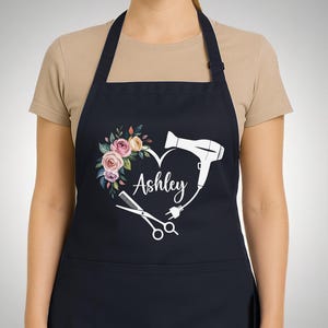 May include: A navy apron with a heart-shaped design, floral elements, and the name "Ashley". The heart includes a hairdryer, scissors, and a comb, indicating a hairstylist theme. The apron is a practical garment.