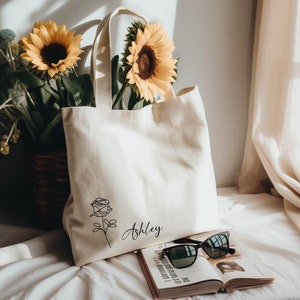 May include: Cream-colored tote bag with a black rose illustration and the name "Ashley" in script. The bag is next to an open book, sunglasses, and sunflowers in a basket. The scene is lit by natural light.