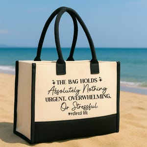 A black and white tote bag with text printed on it, sitting on a sandy beach with the ocean in the background.