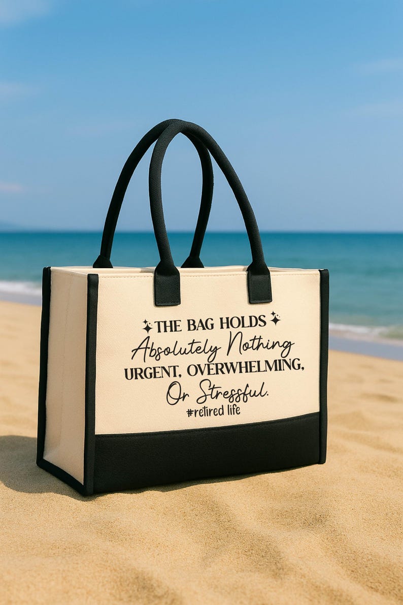 A black and white tote bag with text printed on it, sitting on a sandy beach with the ocean in the background.