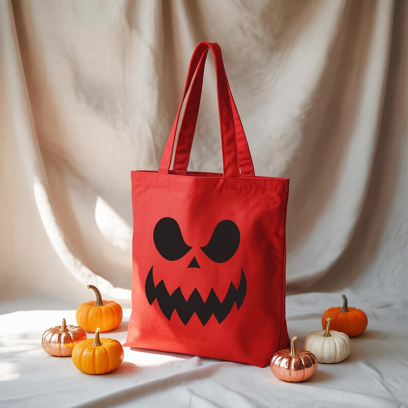 A red tote bag with a Halloween-themed design, including a jack-o-lantern face, is placed on a white surface surrounded by small pumpkins.