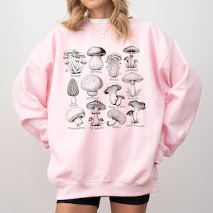 Op de afbeelding: Roze sweatshirt met een grafische afbeelding van verschillende soorten paddenstoelen, waaronder honingzwam, eekhoornzwam, enoki, parasolzwam, stuifzwam, moriel, oesterzwam, spinnenkop, champignon, vliegenzwam, reishi en  zwarte trompetzwam.