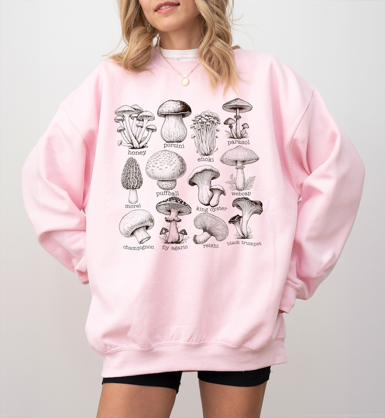 Op de afbeelding: Roze sweatshirt met een grafische afbeelding van verschillende soorten paddenstoelen, waaronder honingzwam, eekhoornzwam, enoki, parasolzwam, stuifzwam, moriel, oesterzwam, spinnenkop, champignon, vliegenzwam, reishi en  zwarte trompetzwam.
