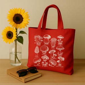 May include: Red canvas tote bag with white illustrations of different types of mushrooms. The bag has two handles and the names of the mushrooms are printed below each illustration. The names include: honey, porcini, enoki, parasol, morel, puffball, king oyster, webcap, champignon, fly agaric, relahi, and black trumpet.
