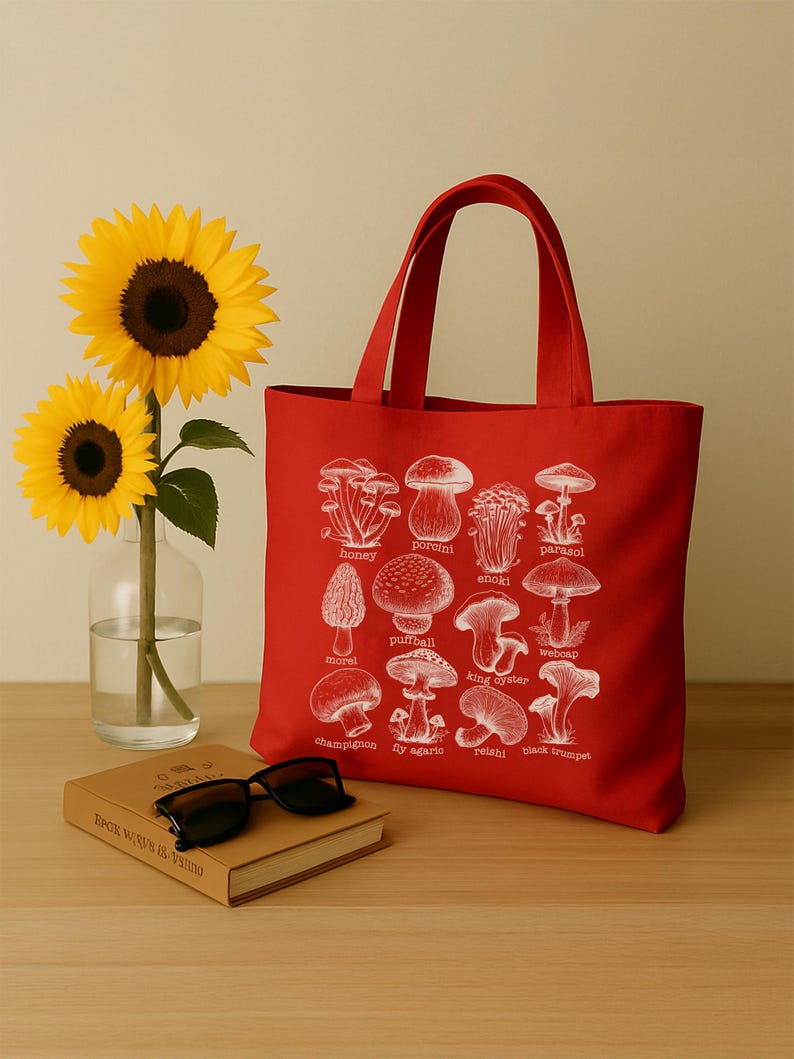 May include: Red canvas tote bag with white illustrations of different types of mushrooms. The bag has two handles and the names of the mushrooms are printed below each illustration. The names include: honey, porcini, enoki, parasol, morel, puffball, king oyster, webcap, champignon, fly agaric, relahi, and black trumpet.