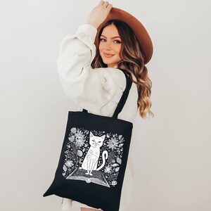 May include: A black tote bag with a white illustration of a cat sitting on a book surrounded by flowers. The bag has long handles.