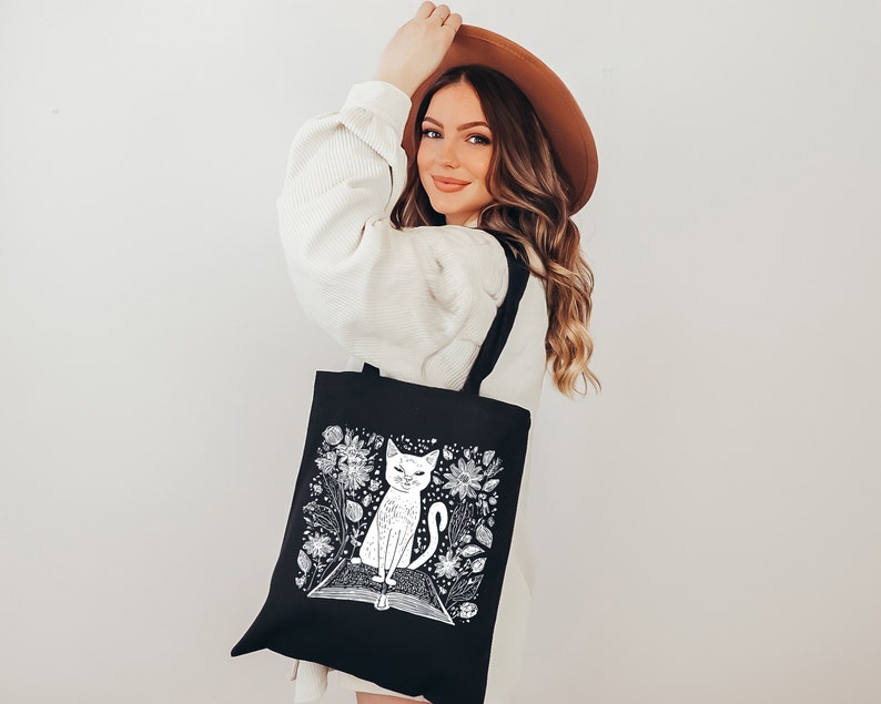May include: A black tote bag with a white illustration of a cat sitting on a book surrounded by flowers. The bag has long handles.