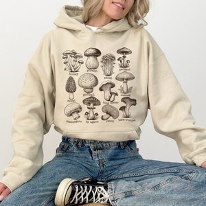 Op de afbeelding: Een beige hoodie met een grafische print van verschillende soorten paddenstoelen met hun namen onder elke afbeelding. De hoodie wordt gedragen door iemand die een blauwe jeans draagt.