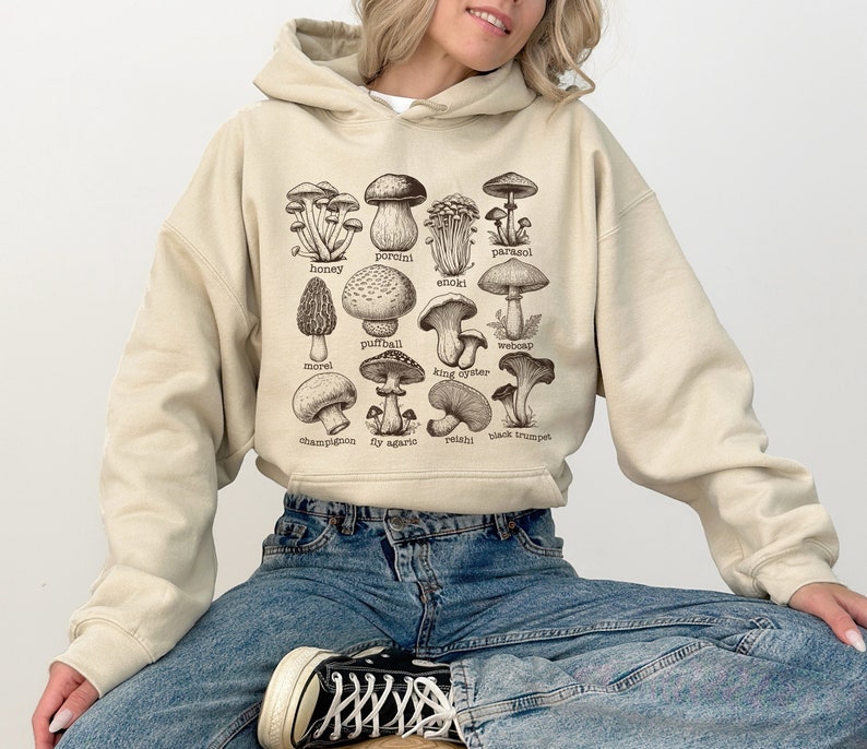 Op de afbeelding: Een beige hoodie met een grafische print van verschillende soorten paddenstoelen met hun namen onder elke afbeelding. De hoodie wordt gedragen door iemand die een blauwe jeans draagt.