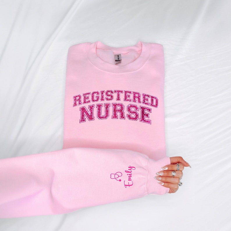 May include: Pink sweatshirt with the text "REGISTERED NURSE" in pink glitter lettering. The left sleeve has a pink embroidered stethoscope and the name "Emily".