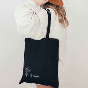 May include: Black tote bag with a white floral design and the name "Isabella" in script. The bag has black straps and is made of a sturdy material. The design is on the lower right corner of the bag.