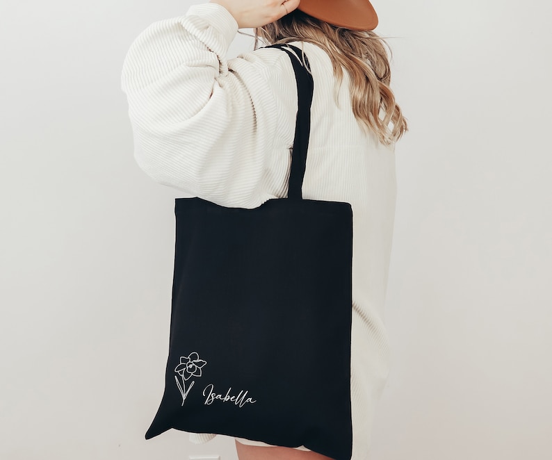May include: Black tote bag with a white floral design and the name "Isabella" in script. The bag has black straps and is made of a sturdy material. The design is on the lower right corner of the bag.
