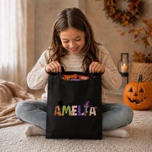 A young girl sits on the floor, holding a black bag with the name "Amelia" printed on it, smiling as she looks inside the bag.