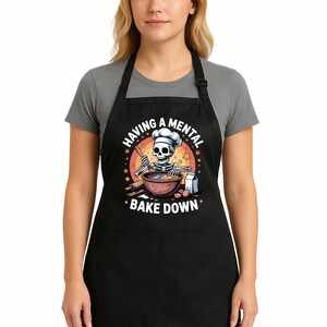 May include: Black apron featuring a cartoon skeleton chef baking. The design shows a skeleton in a chef's hat mixing ingredients, with the text "HAVING A MENTAL BAKE DOWN" in a circular pattern. The apron has a front pocket and is made of a durable fabric.