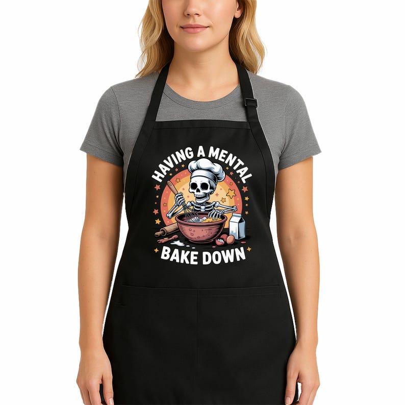 May include: Black apron featuring a cartoon skeleton chef baking. The design shows a skeleton in a chef's hat mixing ingredients, with the text "HAVING A MENTAL BAKE DOWN" in a circular pattern. The apron has a front pocket and is made of a durable fabric.