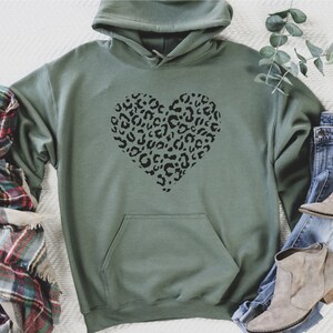 Puede incluir: Una sudadera con capucha verde con un dise&ntilde;o de coraz&oacute;n de estampado de leopardo negro. La sudadera est&aacute; colocada sobre una superficie blanca con una bufanda de cuadros y unos vaqueros con botines marrones.