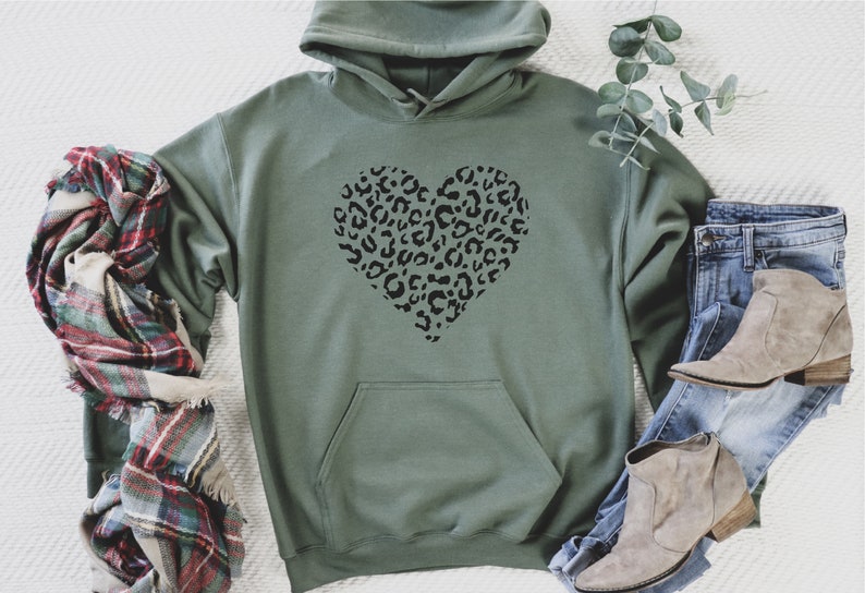 Puede incluir: Una sudadera con capucha verde con un dise&ntilde;o de coraz&oacute;n de estampado de leopardo negro. La sudadera est&aacute; colocada sobre una superficie blanca con una bufanda de cuadros y unos vaqueros con botines marrones.