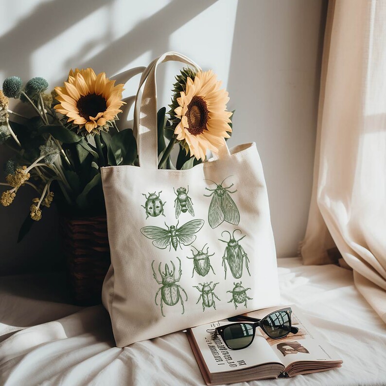 May include: A white canvas tote bag featuring green insect illustrations. The bag has two handles and is resting on a white surface with a book and sunglasses.