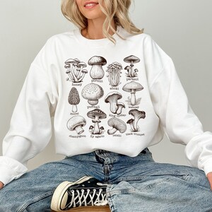 Op de afbeelding: Wit sweatshirt met een grafische print van verschillende soorten paddenstoelen met hun namen onder elke paddenstoel. De namen omvatten: parasol,  eekhoornzwam, honingzwam,  enoki,  stuifzwam,  spinnenkop,  moriel,  oesterzwam,  champignon,  vliegenzwam,  reishi en  trompetzwam.