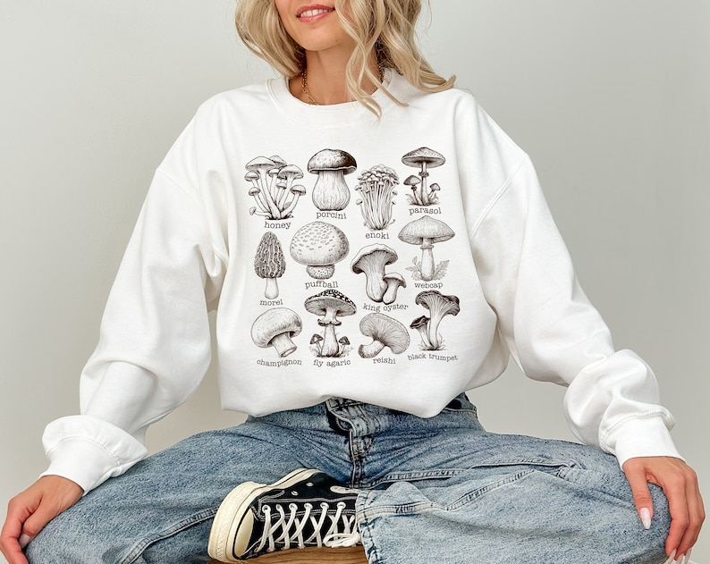 Op de afbeelding: Wit sweatshirt met een grafische print van verschillende soorten paddenstoelen met hun namen onder elke paddenstoel. De namen omvatten: parasol,  eekhoornzwam, honingzwam,  enoki,  stuifzwam,  spinnenkop,  moriel,  oesterzwam,  champignon,  vliegenzwam,  reishi en  trompetzwam.