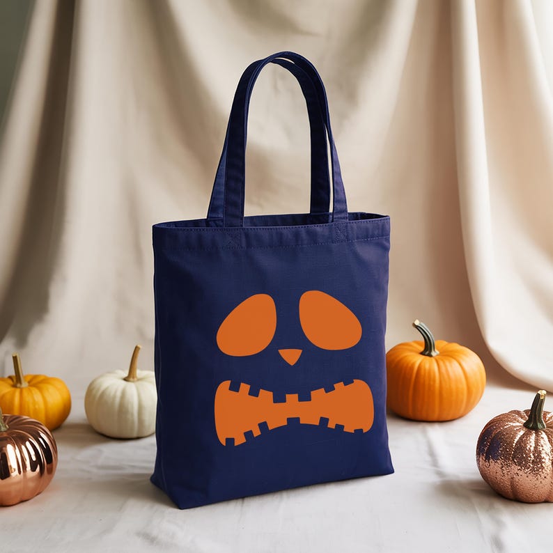A navy blue tote bag with an orange pumpkin face design sits on a white surface, surrounded by four small pumpkins in various colors.