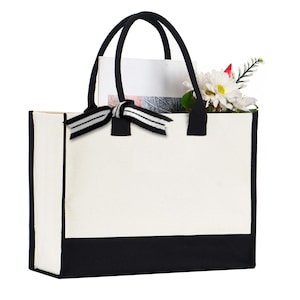 A white tote bag with a black trim, featuring a black bow tied at the top. The bag is holding a bouquet of flowers, including a white daisy and a red flower.